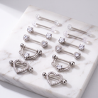 A set of silver-toned body jewelry pieces arranged on a white marble slab, including barbell studs, heart-shaped studs, and a curved ring-style piece with clear rhinestone-like accents.