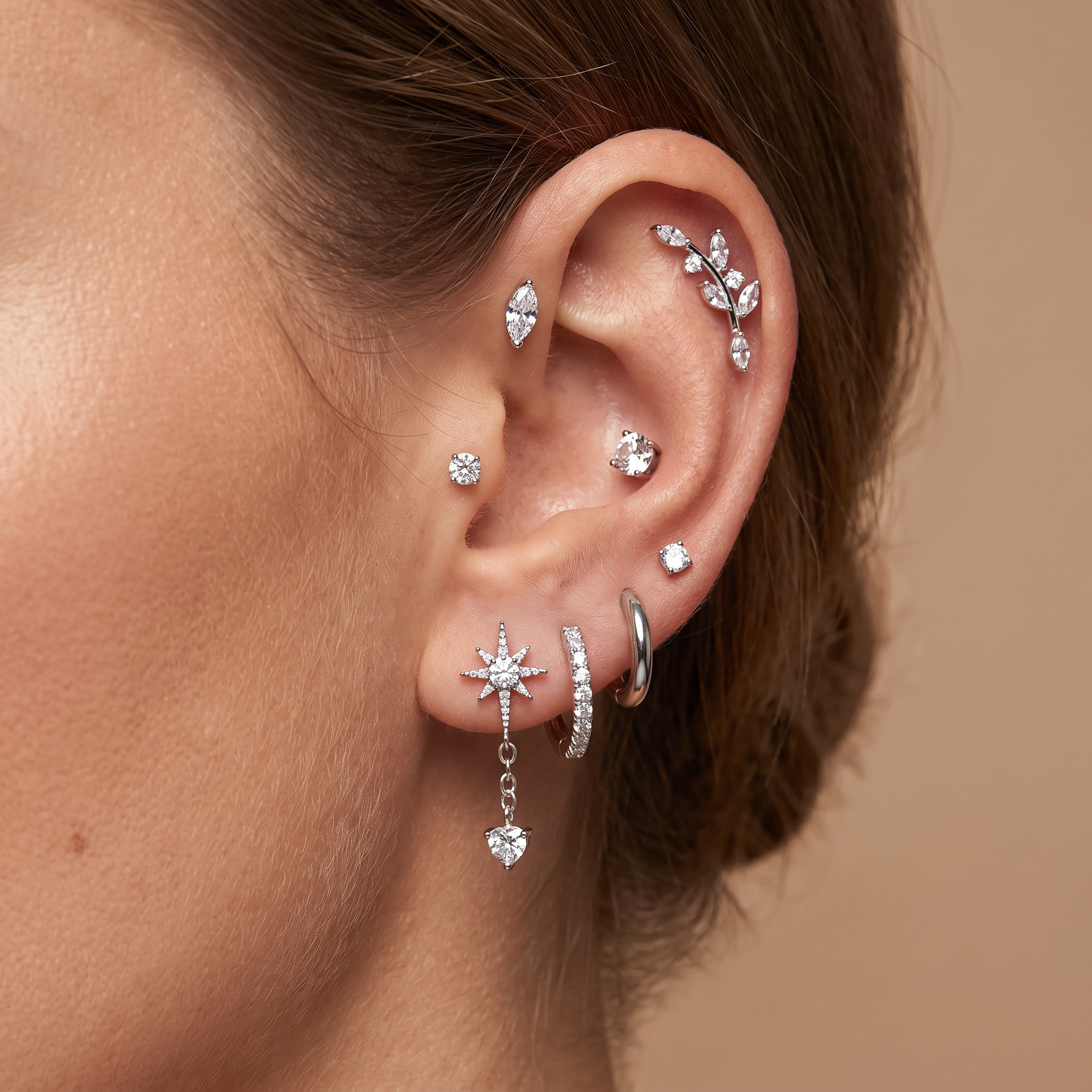 A close-up of a model's ear showcasing a curated collection of silver-toned ear piercings, including multiple studs, a huggie hoop, a leaf cluster cartilage piercing, and a dangling star with a heart charm.