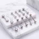Nine silver-toned barbell piercings with clear gemstones are arranged on white marble display blocks against a white marble surface.