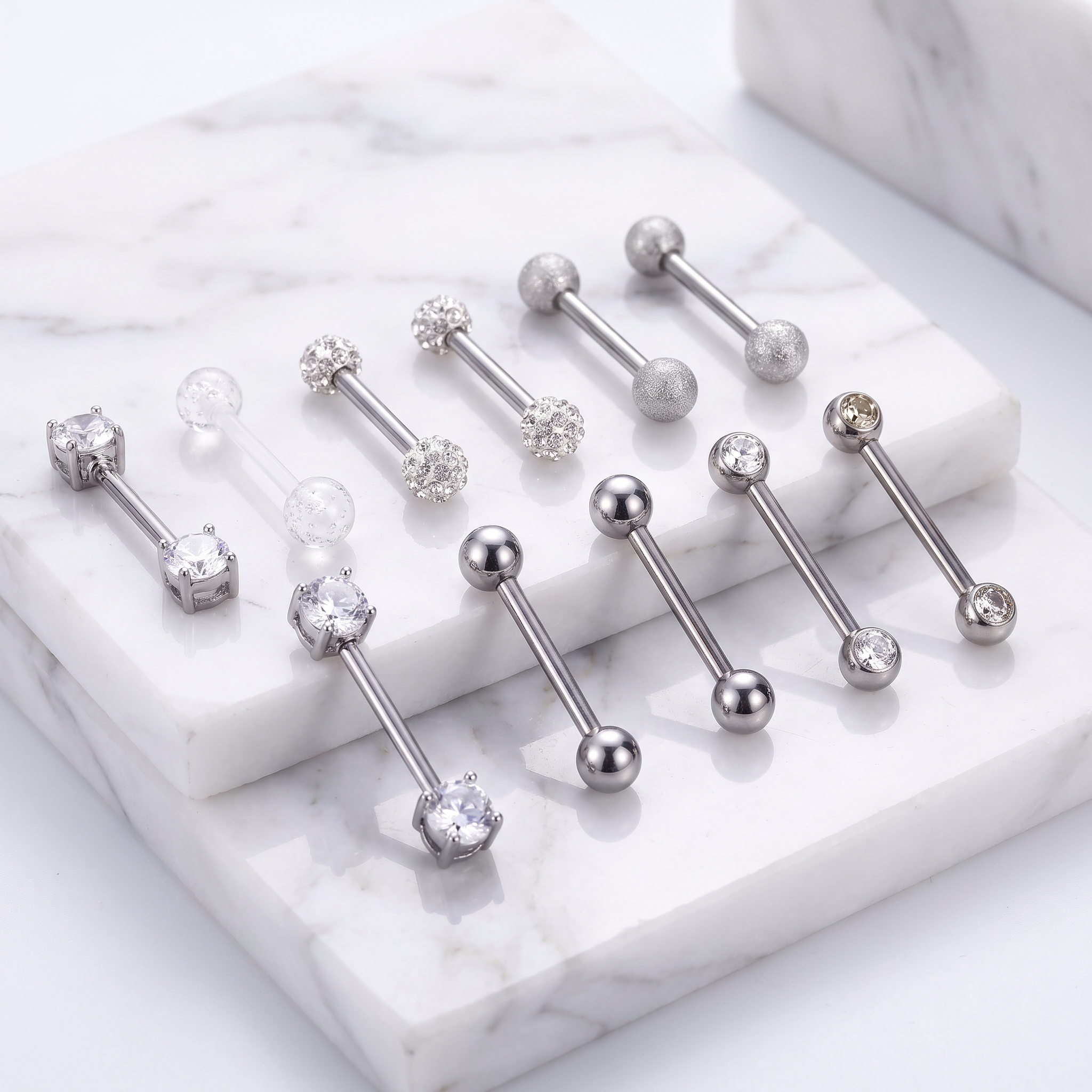 Nine silver-toned barbell piercings with clear gemstones are arranged on white marble display blocks against a white marble surface.
