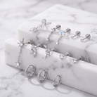 A styled product shot of multiple silver-toned ear piercings and rings displayed on white marble blocks over a marble surface. The jewelry includes hoop earrings, studs, and dangling charms with clear stones in floral, starburst, and marquise shapes.
