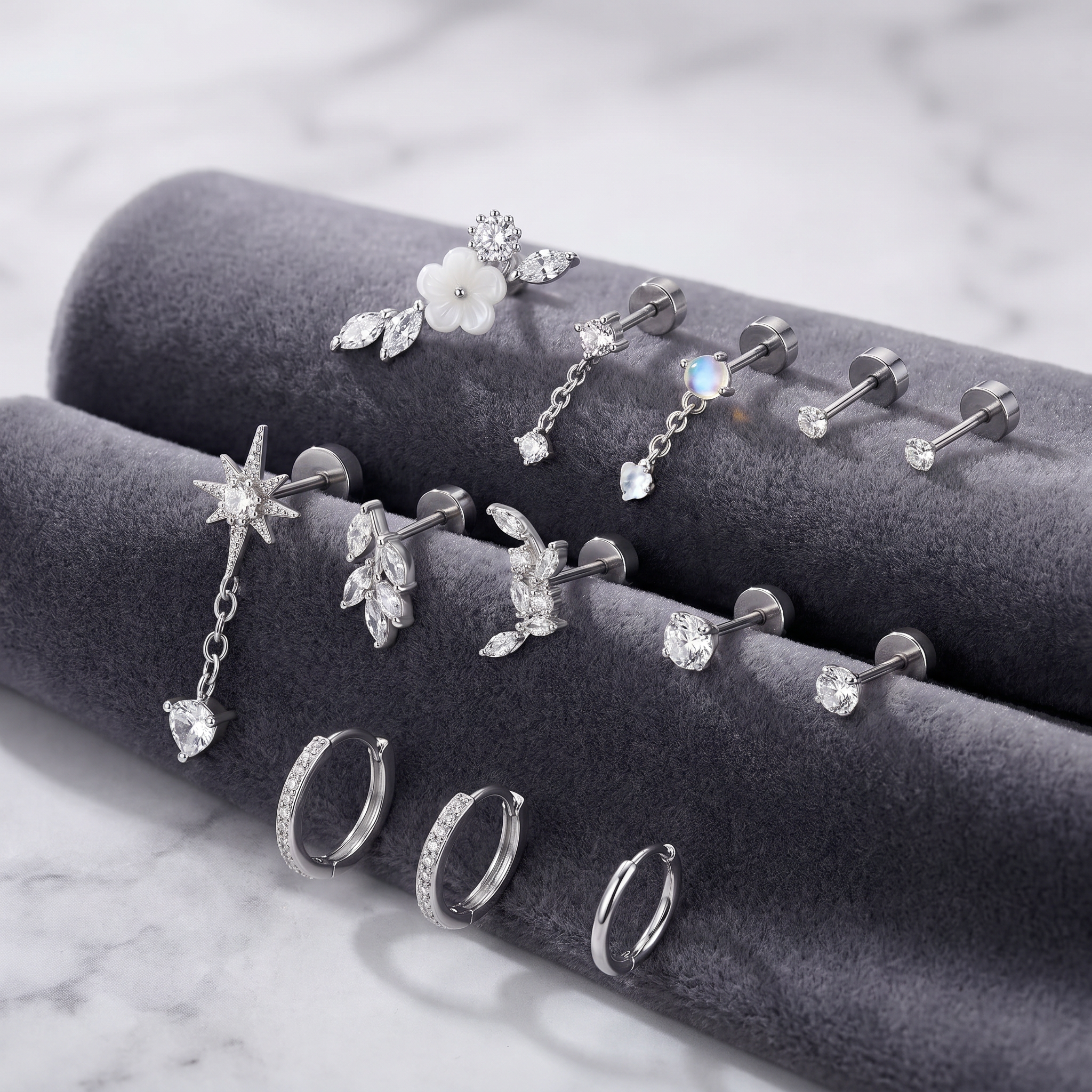 A set of silver-toned body jewelry displayed on dark gray velvet rings, including studs, earrings, and a flower-shaped stud, photographed on a light marble surface.