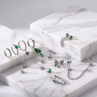 A styled flat-lay of silver-toned body jewelry and earrings with green gemstones, arranged on white marble display blocks against a light gray background.