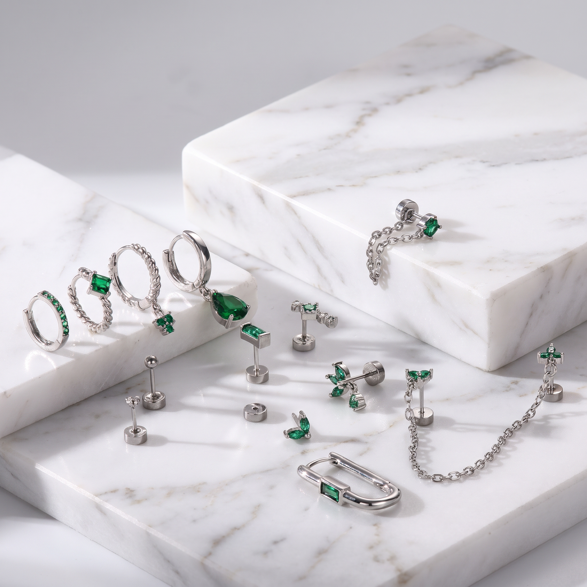 A styled flat-lay of silver-toned body jewelry and earrings with green gemstones, arranged on white marble display blocks against a light gray background.