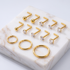 Gold body jewelry set displayed on a white marble slab, featuring multiple straight barbells with clear gemstones and three matching curved hoop rings.