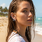 A woman on a sandy beach wearing multiple silver-toned ear cuff piercings and dangling earrings, with ocean waves and palm trees in the background.