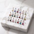 A set of colorful belly button piercings arranged on a white marble tray, featuring silver-toned bars with round gemstone tops in a variety of colors.