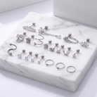 A set of silver-toned body jewelry pieces arranged on a white marble display block, including studs, rings, and curved barbell-style earrings with clear stones and metallic charms.