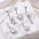 Five silver-toned belly button rings with clear rhinestones and crystal-like gemstones, arranged on a white marble block.