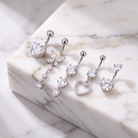 Four silver-toned belly button rings with clear crystal charms, including a heart, butterfly, triple-heart drop, and heart-with-stones design, arranged on a white marble slab.