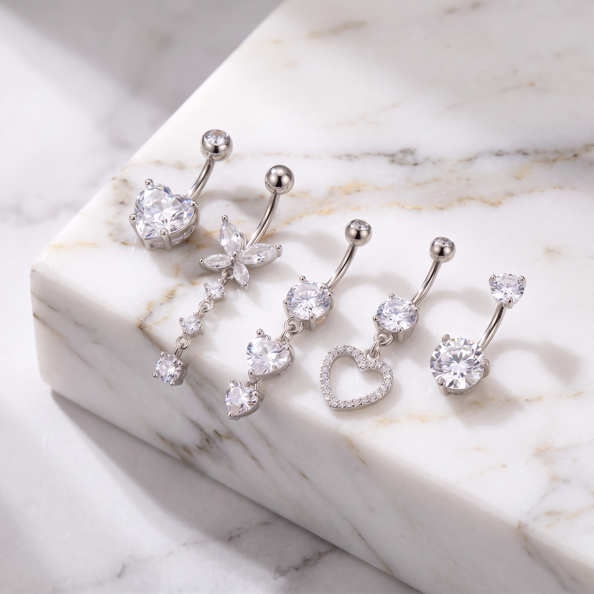 Four silver-toned belly button rings with clear crystal charms, including a heart, butterfly, triple-heart drop, and heart-with-stones design, arranged on a white marble slab.