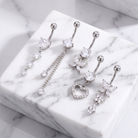 Four silver-toned belly button rings with clear rhinestone and crystal charms, arranged on a white marble block against a light marble surface.