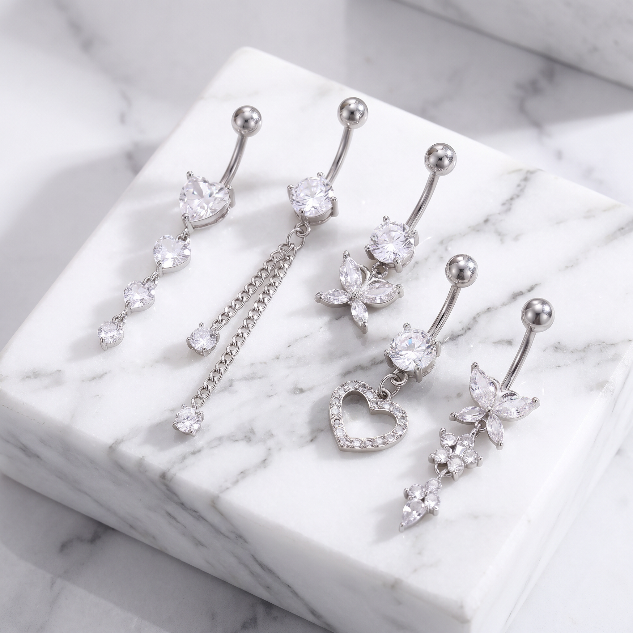 Four silver-toned belly button rings with clear rhinestone and crystal charms, arranged on a white marble block against a light marble surface.