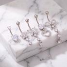 Five silver-toned belly button rings with clear rhinestone and crystal charms, displayed on a white marble block and marble surface.
