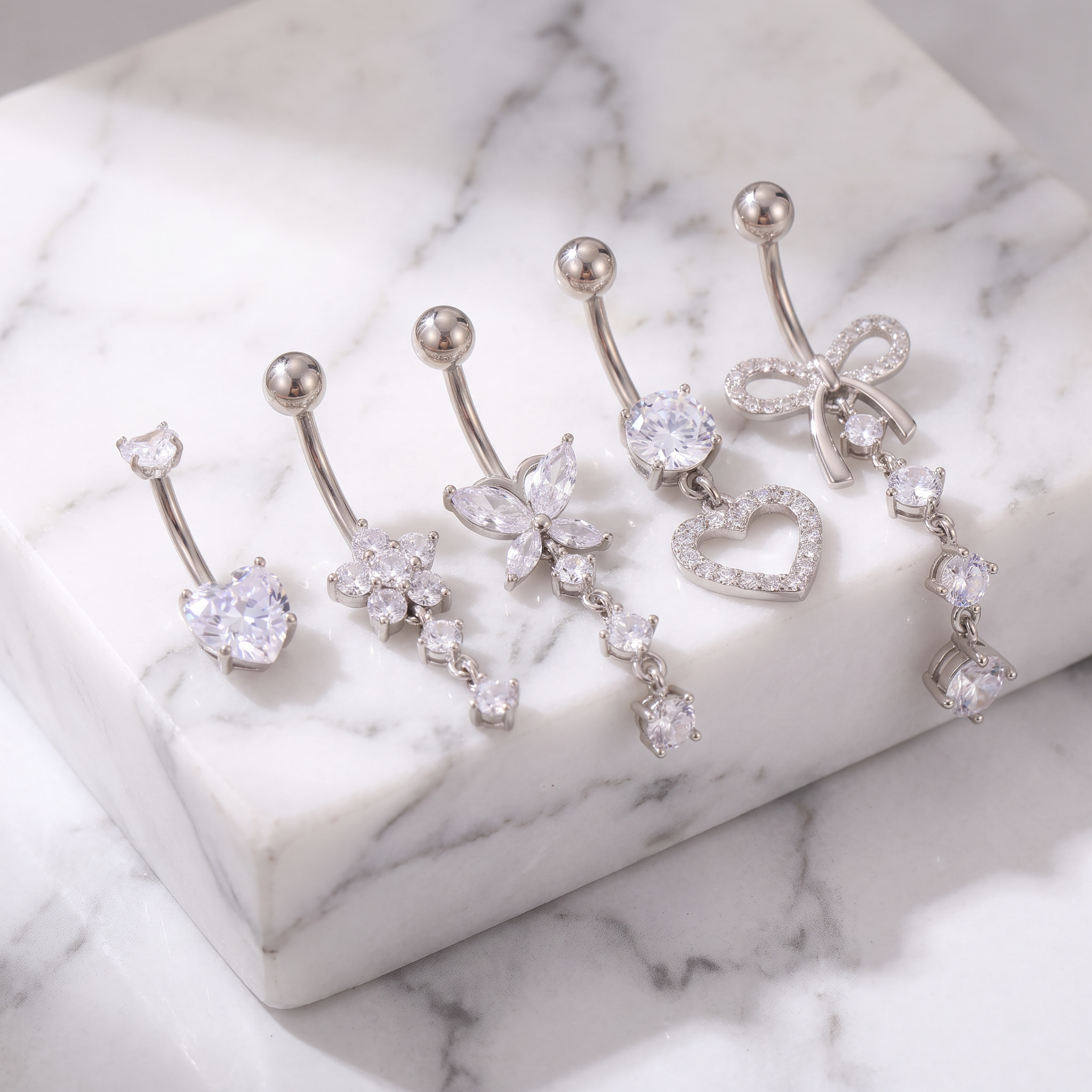 Five silver-toned belly button rings with clear rhinestone and crystal charms, displayed on a white marble block and marble surface.