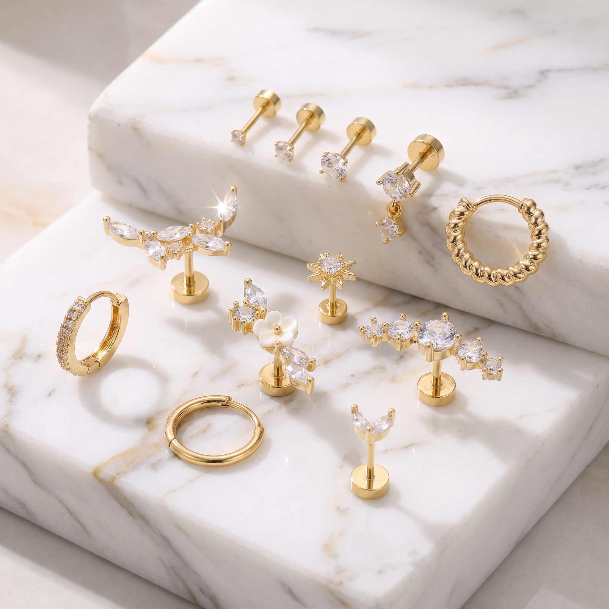 A set of gold-toned body jewelry and hoop earrings displayed on white marble blocks. The collection includes flat-back labret studs, hoop earrings, butterfly-shaped studs, flower-shaped studs, and a textured rope hoop.