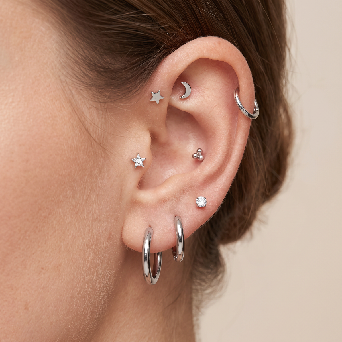 Close-up of a person's ear adorned with multiple silver piercings, including hoop earrings, a crescent moon stud, a star stud, a small three-stone stud, and a flat circle stud, against a soft beige background.