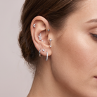 A close-up of a model's ear showcasing multiple silver-toned ear piercings, including a crescent moon stud, a cluster of small floral studs, a dangling star charm, a huggie hoop, and a pavé huggie hoop, against a soft beige background.