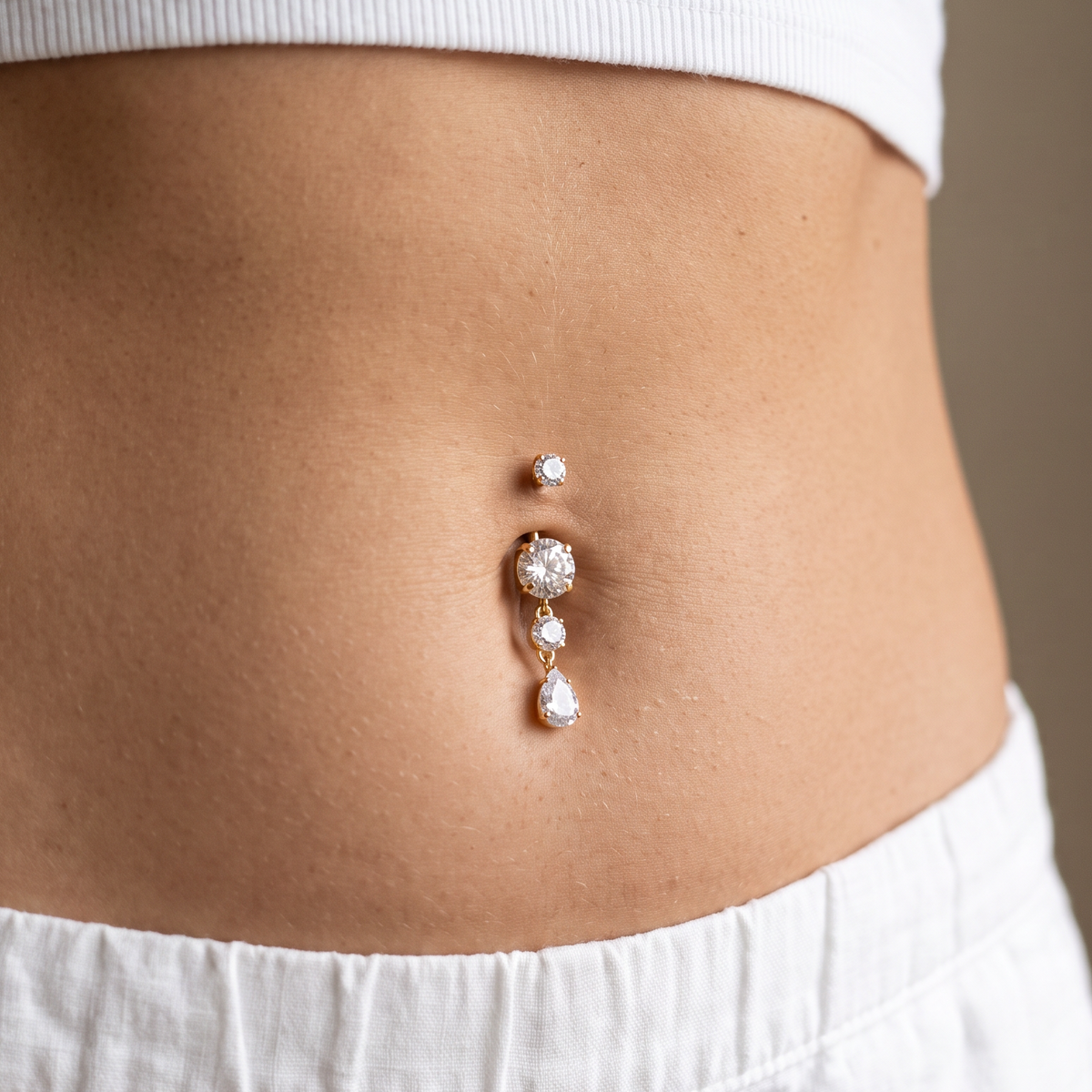 A close-up of a person's lower torso wearing a silver belly button ring with three clear rhinestone-like drops, styled with a white ribbed crop top and white pants.