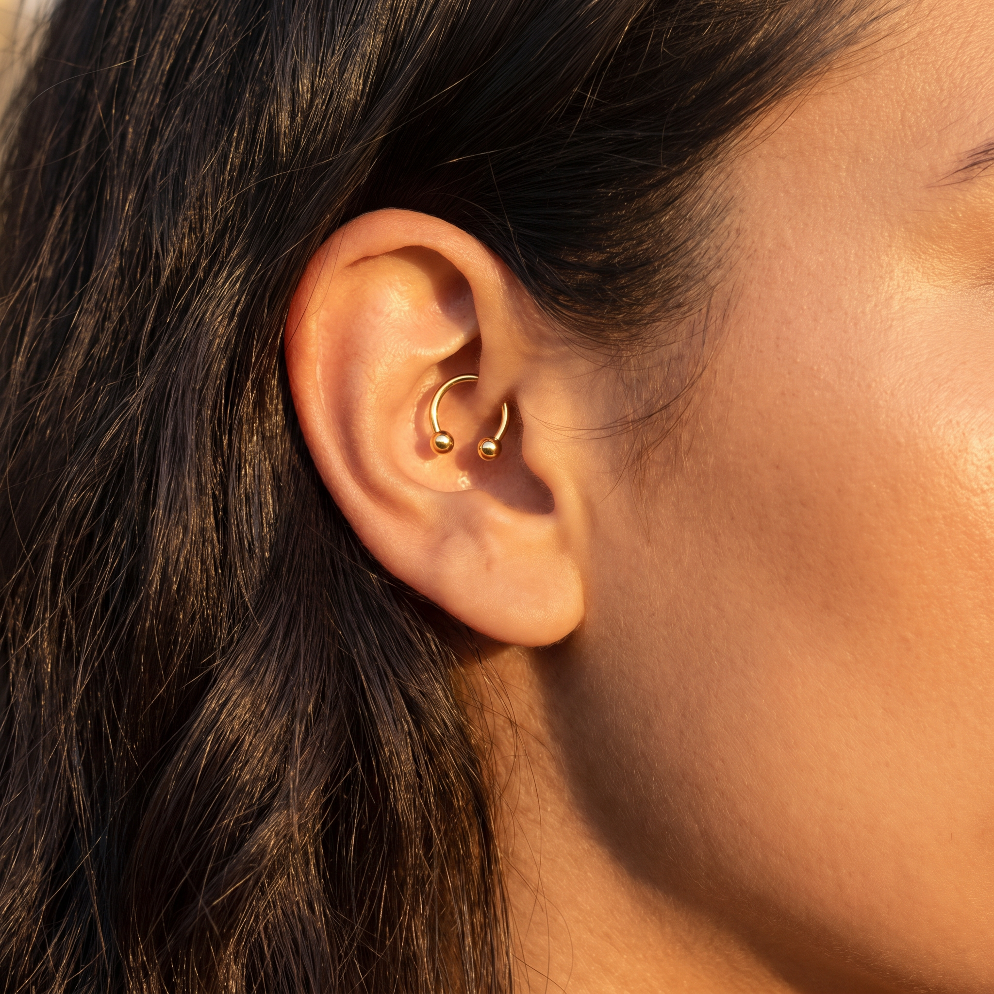 A close-up of a person's ear wearing a small gold hoop piercing in the cartilage and a tiny gold ball stud above it, with dark hair and warm sunlight.