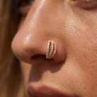 A close-up of a person's nose wearing a gold double-row nose stud with small clear stones, set against warm natural sunlight.
