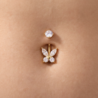 A close-up of a belly button piercing with a gold butterfly-shaped stud adorned with clear marquise and round gemstones, shown on skin.