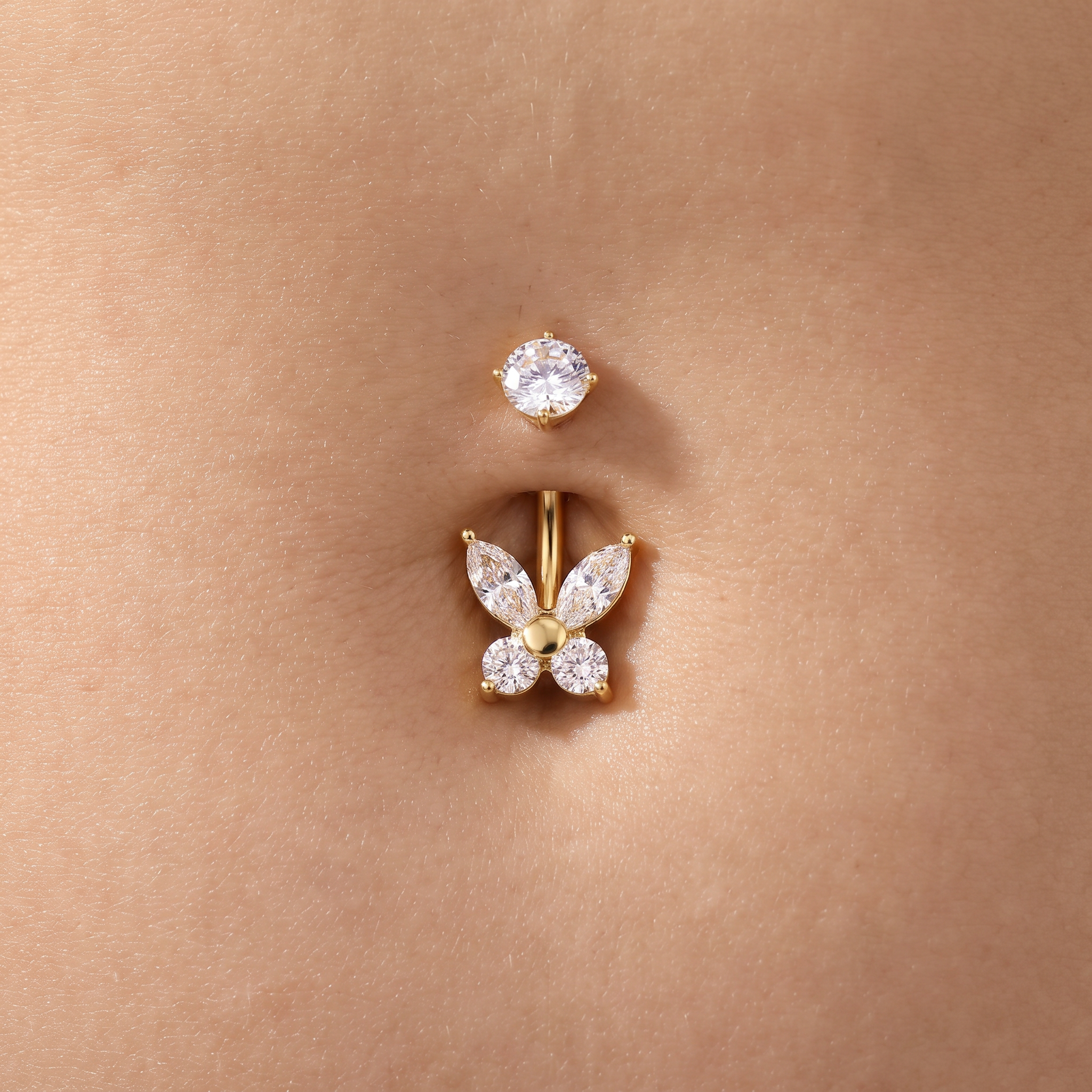 A close-up of a belly button piercing with a gold butterfly-shaped stud adorned with clear marquise and round gemstones, shown on skin.