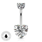 A silver-toned belly button ring with a heart-shaped clear gemstone at the bottom and a smaller heart-shaped gemstone at the top, shown on a white background with a circular skin-safe waterproof badge in the lower left.