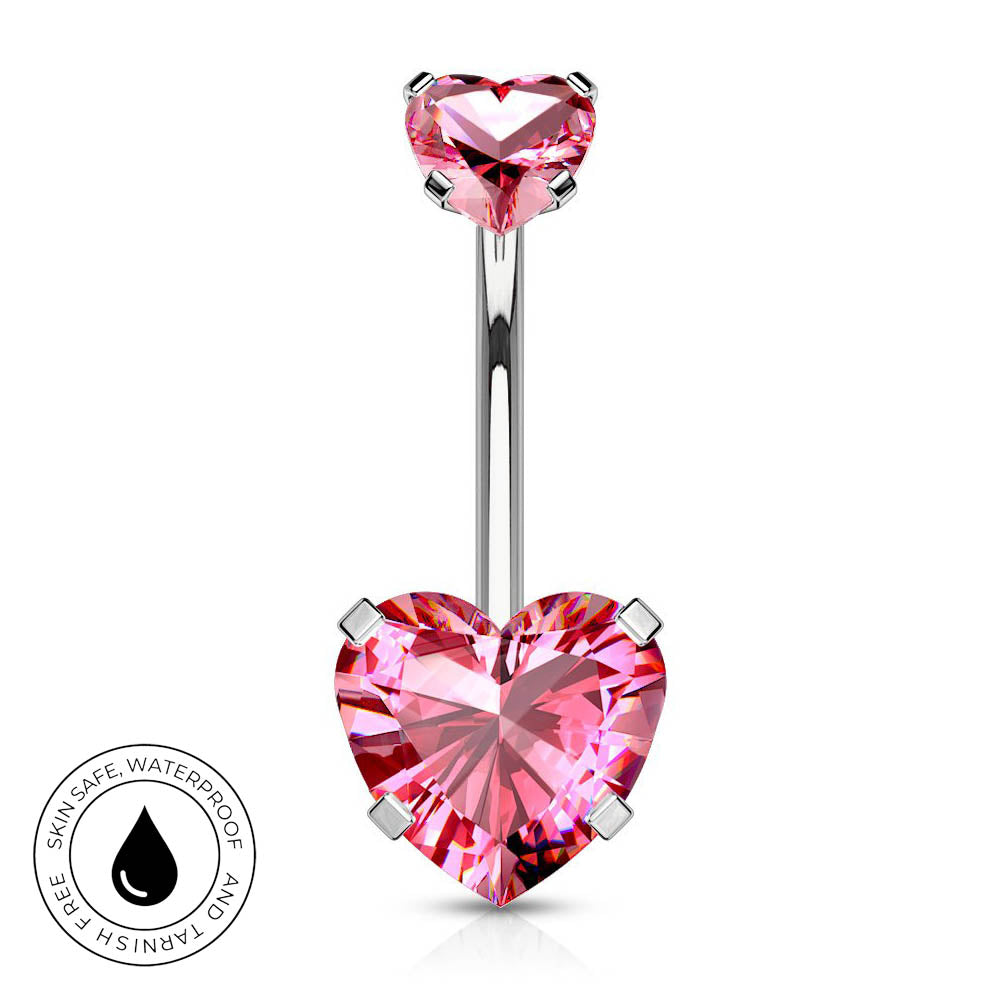 A silver-toned navel barbell piercing with a large, faceted pink heart-shaped crystal at the bottom and a smaller matching pink heart at the top. A circular badge in the bottom-left corner highlights skin-safe, waterproof quality.