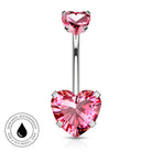 A silver-toned navel barbell piercing with a large, faceted pink heart-shaped crystal at the bottom and a smaller matching pink heart at the top. A circular badge in the bottom-left corner highlights skin-safe, waterproof quality.