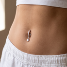 A close-up of a person's midsection wearing a silver crescent-shaped navel piercing with a dangling teardrop crystal, styled with a white ribbed crop top and white shorts.