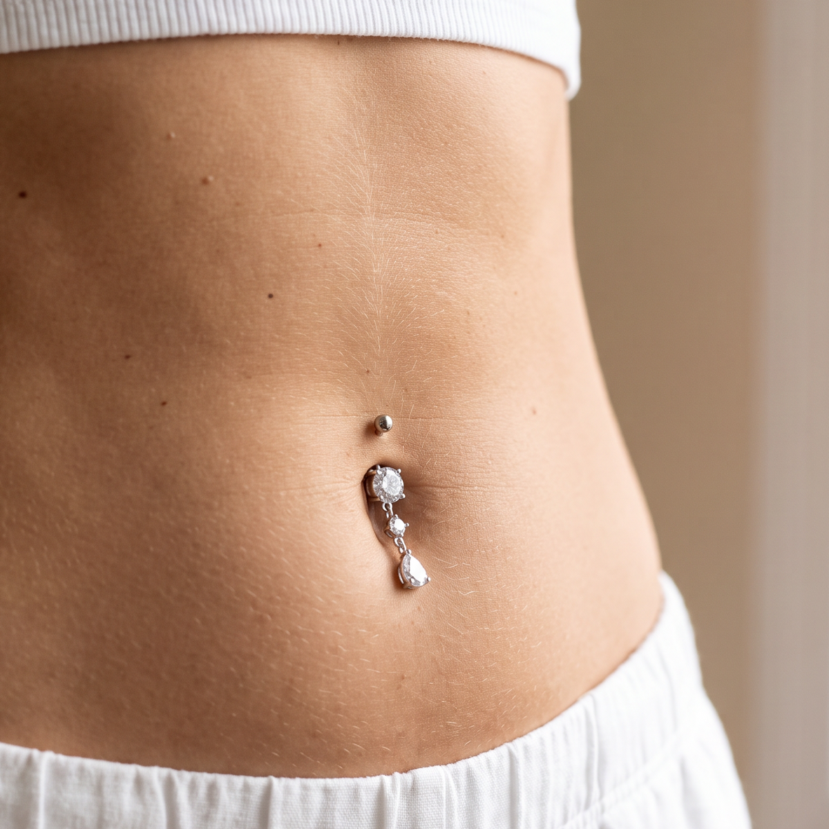 A close-up of a person's midsection wearing a silver belly button ring with clear rhinestones, paired with a white ribbed crop top and white pants.