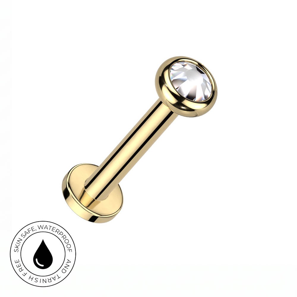 A gold-plated labret piercing barbell with a clear faceted gemstone in the end, shown on a plain white background with a circular 'Skin Safe Waterproof' badge in the bottom-left corner.