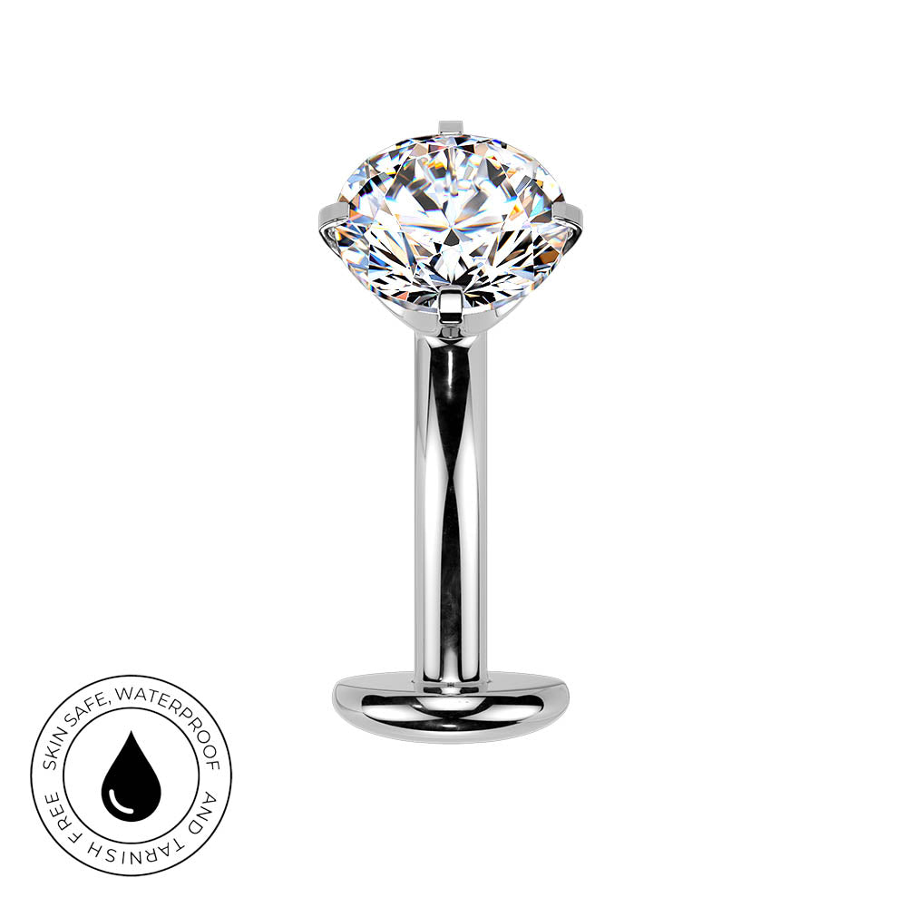 A polished silver-toned belly piercing bar with a large clear faceted gemstone, shown on a plain white background. A circular badge in the lower-left corner reads 'SKIN SAFE WATERPROOF AND TARNISH FREE' with a droplet icon.