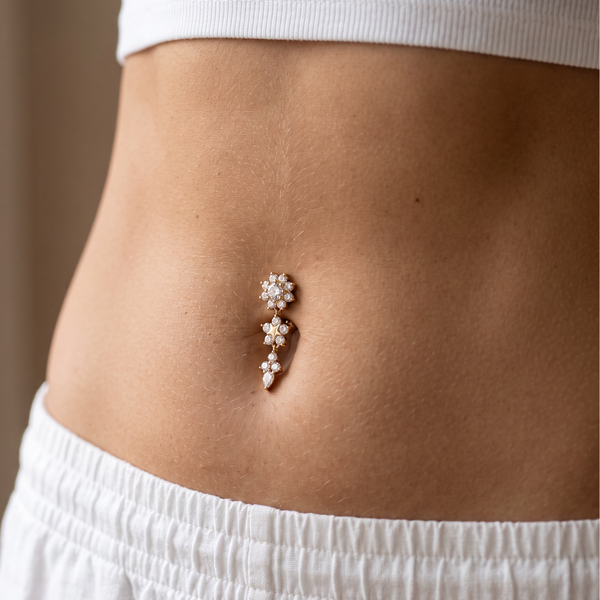 A close-up of a person's lower torso wearing a small belly button ring with three dangling flower clusters in a rose gold-toned metal and clear stones.