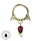 Gold-toned piercing ring with curved claw-like prongs and a dangling red teardrop gemstone, shown on a white background with a circular skin-safe waterproof badge in the lower left.