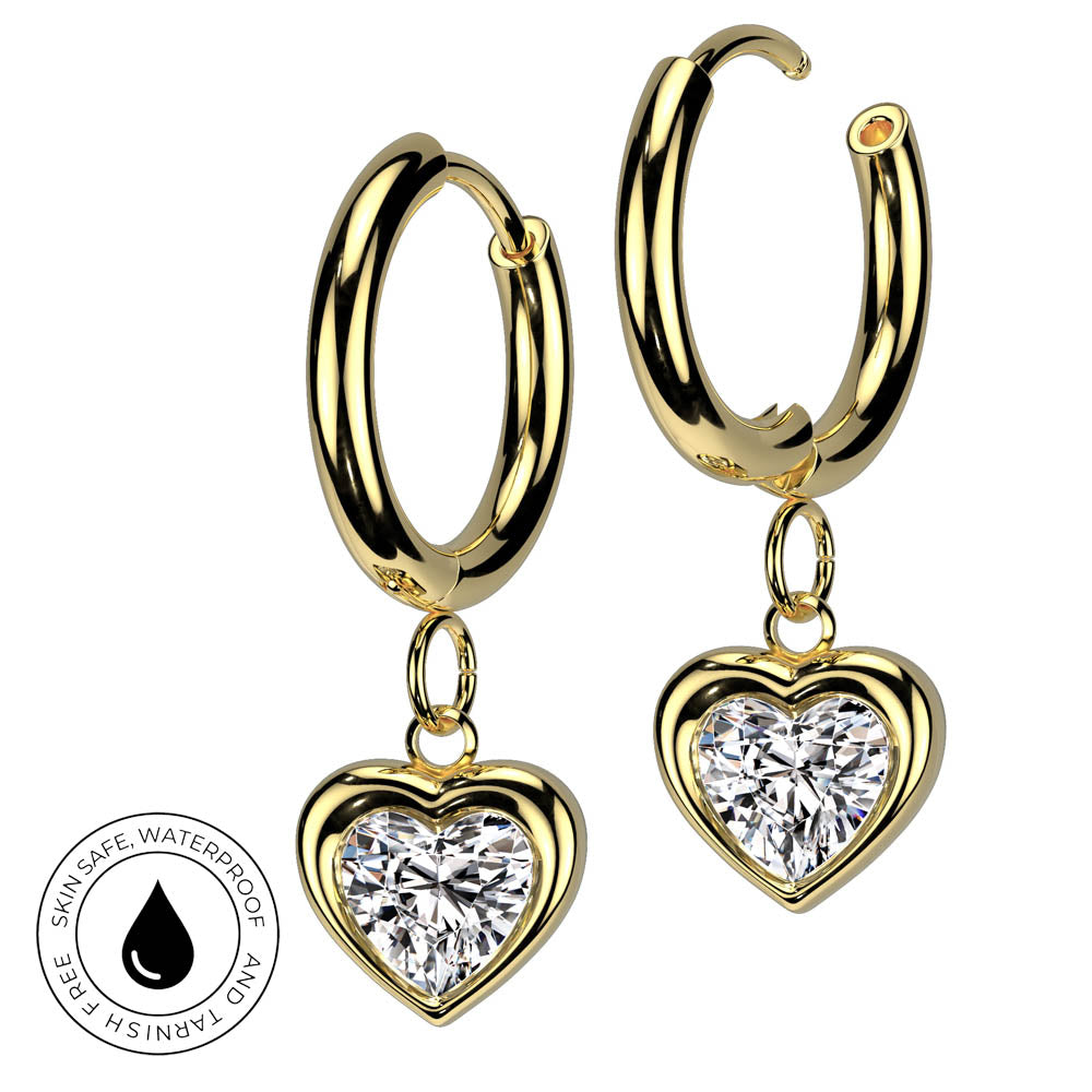 A pair of gold hoop earrings with dangling heart-shaped settings holding clear heart-shaped gemstones, shown on a white background with a small circular skin-safe waterproof badge in the lower left.
