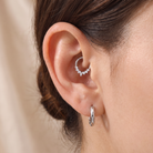 A close-up of a person's ear showcasing a silver crescent hoop earring in the cartilage with small clear stones, paired with a classic silver hoop earring below, against a soft beige background.