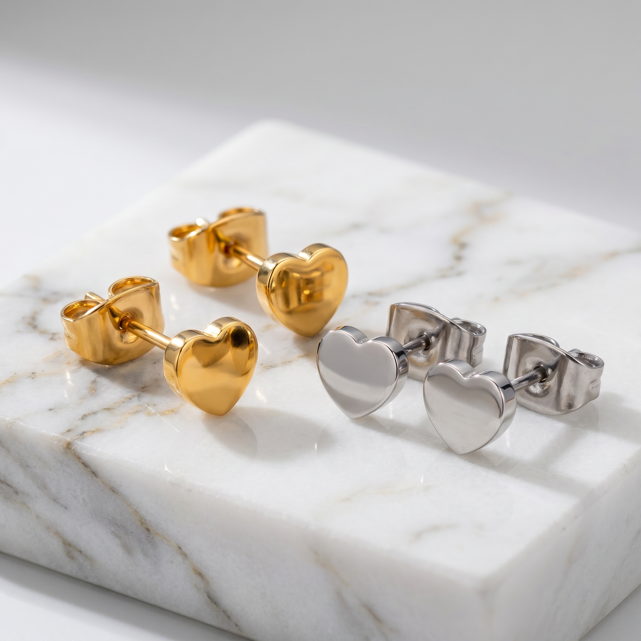 Two pairs of heart-shaped stud earrings, one in polished yellow gold and one in polished silver, displayed on a white marble block.