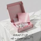 An open pink gift box labeled CHERRY DIVA sits on a white marble surface, filled with pink crinkle paper and a small clear pouch containing silver-toned jewelry. A matching pink card is placed in front, and text at the bottom reads 'WRAP IT UP with our bespoke gift box.