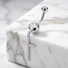 A silver-toned navel piercing ring with a curved barbell, a round bezel-set clear crystal, and a dangling cross charm adorned with small clear crystals, displayed on a white marble block.