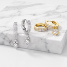 A set of gold and silver huggie hoop earrings embellished with pavé stones and dangling clear crystal charms, displayed on a white marble block.